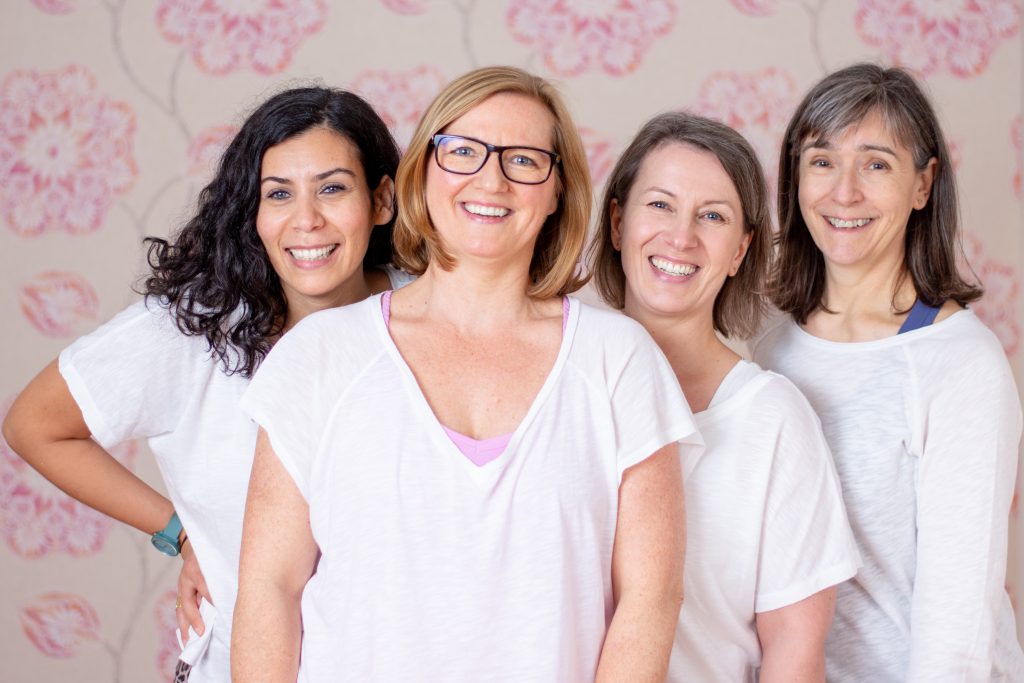 Vier lächelnde Frauen in weißen Shirts stehen vor einer hellen, geblümten Wand und blicken freundlich in die Kamera. Gruppenfoto für Business- oder Teampräsentation.