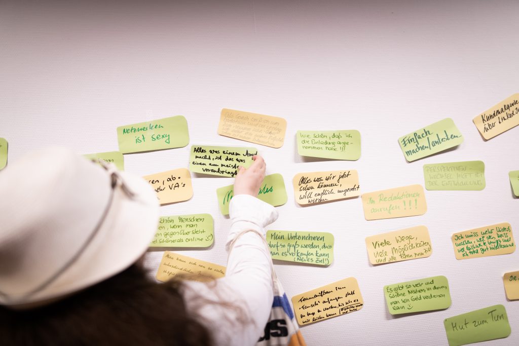 Person in a white hat reaching towards colorful sticky notes on a wall, each with handwritten notes in German.