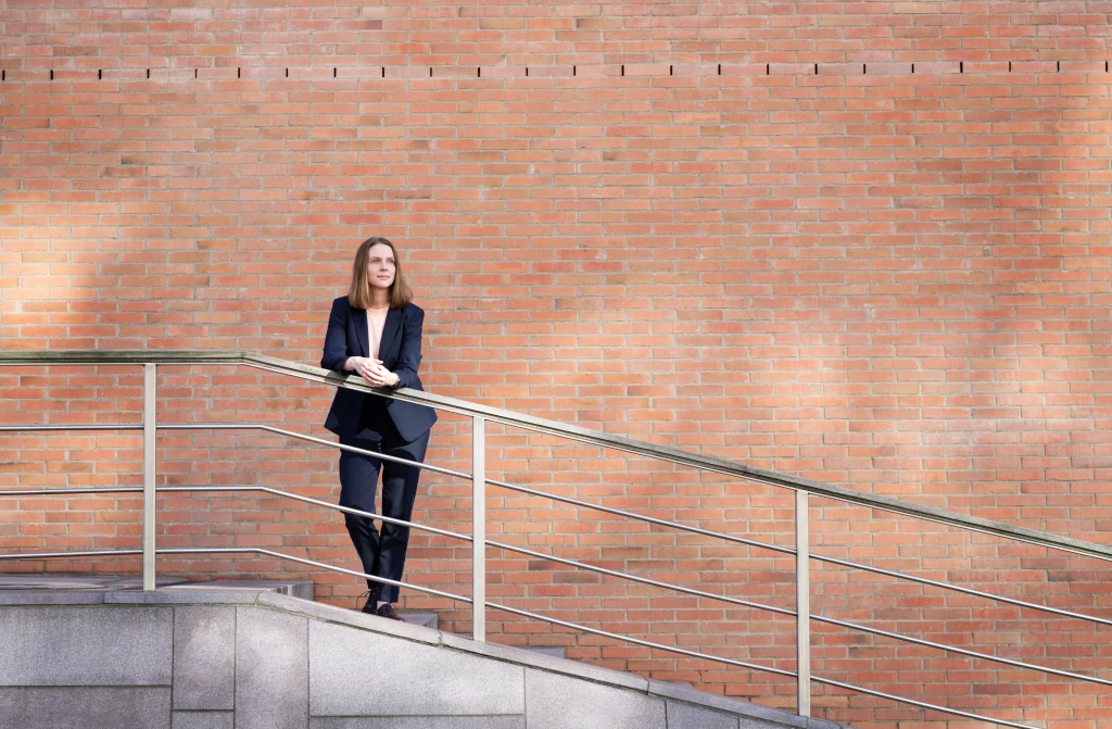 Frau im Business-Outfit steht draußen an einem Geländer vor einer großen roten Backsteinwand, modernes Businessportrait mit viel Raum.